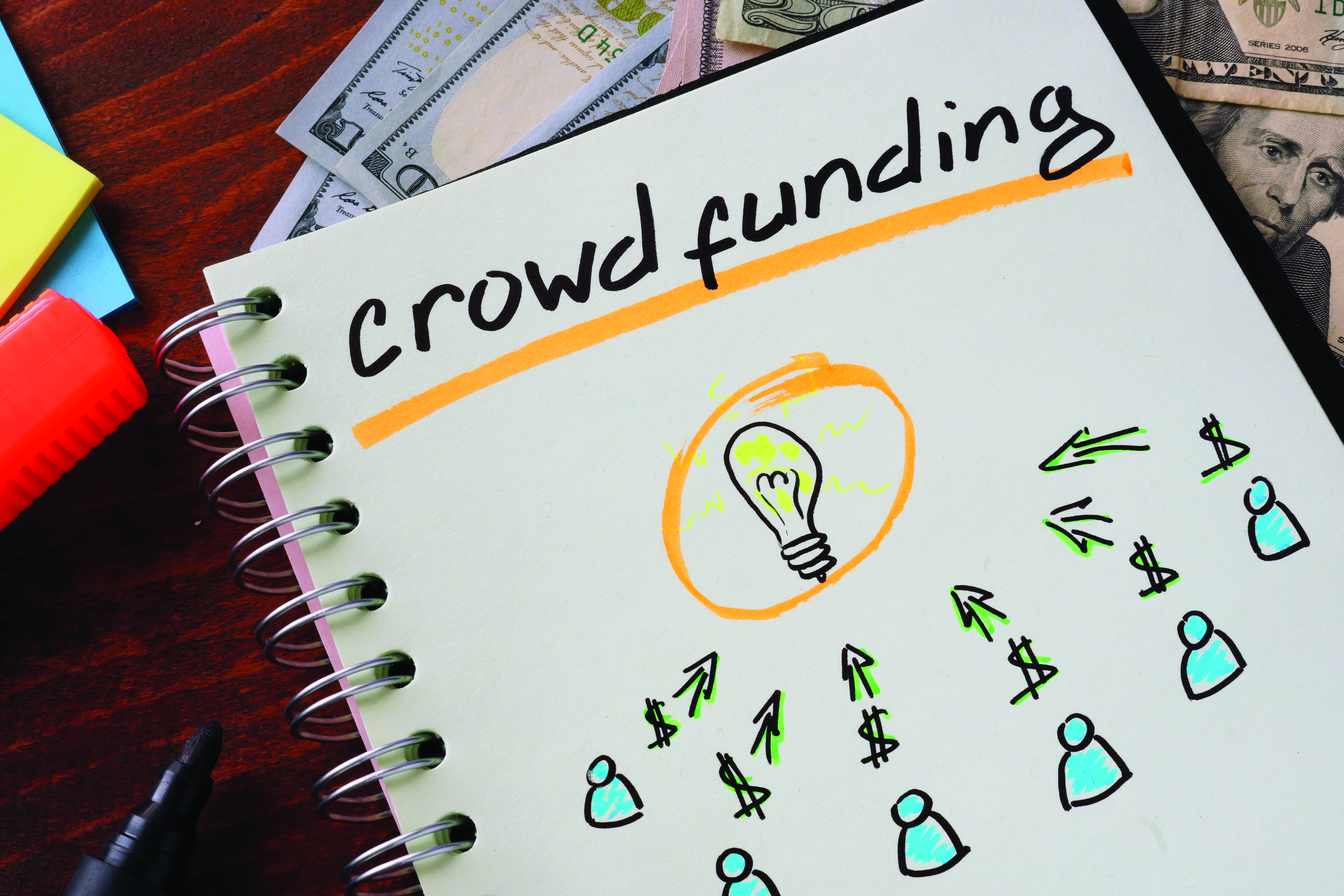 On a notebook, a drawing of people sending money towards a lightbulb is depicted to represent crowdfunding an idea for a business.